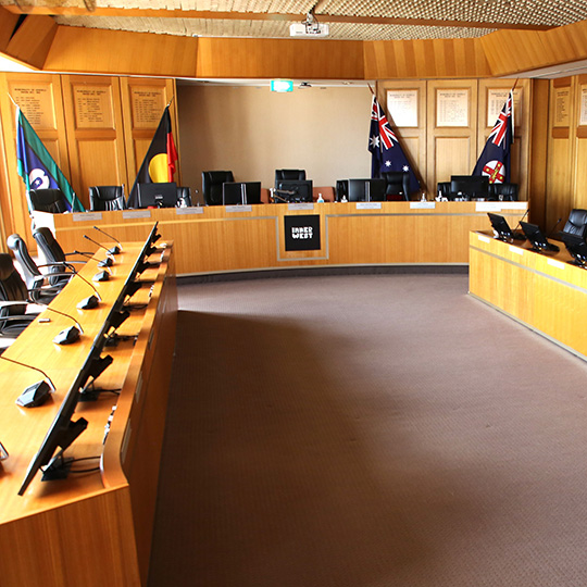 A wood-panelled council chambers with three long desks - equipped with monitors and microphones - arranged in a U shape. The 3 official Australian flags and the New South Wales flag are positioned behind the front desk.